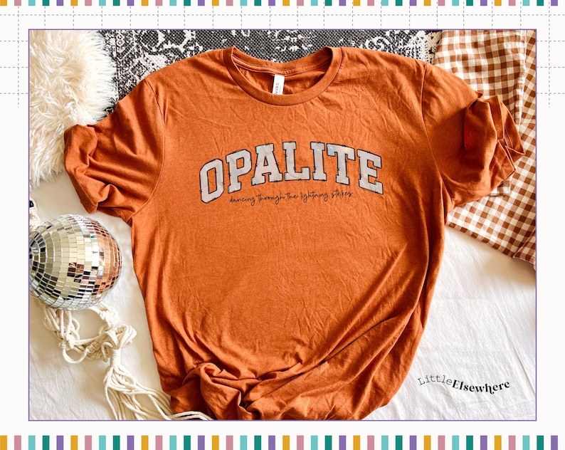 May include: A burnt orange short-sleeve t-shirt featuring the word OPALITE in large, distressed white block letters with a black outline, and the cursive text dancing through the lightning strikes printed underneath the main graphic.