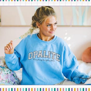 May include: A light blue crewneck sweatshirt featuring the word OPALITE in large white block letters with a dark outline, and the smaller text dancing through the lightning strikes printed underneath in a black cursive script font.