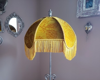May include: A gold lampshade with a fringe trim and a floral pattern. The lampshade is mounted on a silver metal stand.