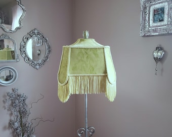 May include: A green lampshade with a fringe trim. The lampshade is made of a soft, textured fabric and has a floral pattern. The lampshade is mounted on a tall, slender lamp stand.