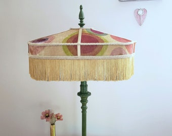 May include: A green table lamp with a round, fringed lampshade. The lampshade is made of a cream-colored fabric with a pattern of pink, green, and red circles. The lampshade has a white trim and a gold fringe.