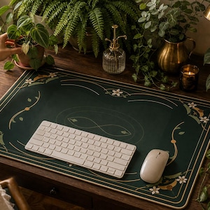 May include: A rectangular dark green desk mat featuring a decorative gold border with white floral accents and an infinity symbol in the center. A white wireless keyboard and a white computer mouse are placed on top of the mat on a wooden desk.