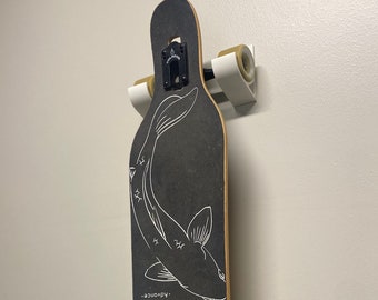 Puede incluir: Una tabla de skate larga negra con un gráfico blanco de un pez en la cubierta. La tabla de skate está montada en un soporte de pared blanco con ruedas doradas. El texto "Valador - Advance" está impreso en la cubierta.