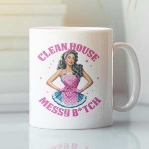 Op de afbeelding: Witte keramische mok met een roze en blauwe illustratie van een vrouw in een roze en witte stippen schort. De tekst "Clean House Messy B*tch" is in roze op de mok gedrukt.