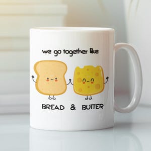 Può includere: Una tazza in ceramica bianca con un'illustrazione in bianco e nero di una fetta di pane e un pezzo di burro con facce. Il testo "We go together like bread & butter" è stampato sulla tazza.