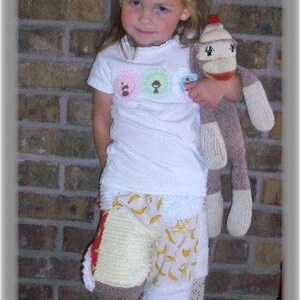 May include: A young girl wearing a white shirt with colorful circles and a patchwork skirt. She is holding a brown and white monkey sock puppet. The girl is wearing a red and black bow in her hair.