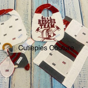 May include: A collection of baby items, including bibs, a blanket, and a teether. The bibs feature a red and white design with the text "City Rescue Team" and a firefighter's helmet. The blanket has a patchwork design.