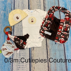 May include: A baby gift set featuring a white onesie with a monkey applique, a yellow striped hat, a bib, and a wooden ring toy. The bib and toy have a monkey sock print. The set is for 0/3m babies.