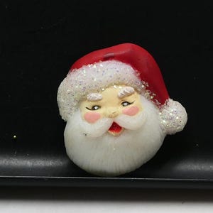 Pode incluir: Uma figura de cabeça de Pai Natal com um gorro vermelho com uma borda e um pompom brancos brilhantes. O rosto tem barba, bigode e sobrancelhas brancas, com bochechas rosadas e uma pequena boca aberta que mostra lábios de cor vermelha.