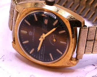 Op de afbeelding: Een gouden polshorloge met een bruine wijzerplaat en een gouden metalen band. De wijzerplaat van het horloge heeft een dag- en datumaanduiding, en de tekst "LEON PIRADET" en "21 JEWELS" staat erop gedrukt.