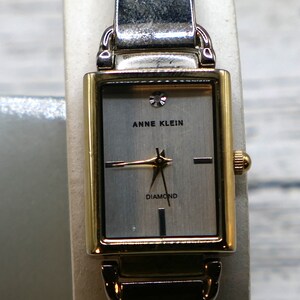 May include: A gold and silver two-tone watch with a rectangular face. The watch face is white with black hands and a diamond at the 12 o'clock position. The watch face also has the text "Anne Klein" and "Diamond".