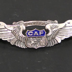 May include: A silver-coloured military-style pin with wings, featuring a blue and silver emblem with the letters "CAF" and the text "GHOST SQUADRON". The pin includes the years 1939 and 1945.