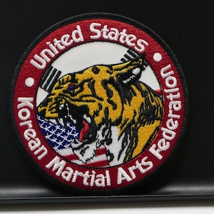 Può includere: Toppa rotonda con motivo a tigre. La toppa presenta il testo "United States Korean Martial Arts Federation" in una disposizione circolare attorno alla tigre. La tigre è gialla, nera e rossa, con una bandiera blu e bianca in bocca.