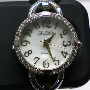 May include: A silver-tone analog wristwatch featuring a round white dial with dark numerals, the text STUDIO and QUART, and gold-tone hands. The bezel is encrusted with small clear crystals, and the watch case connects to a silver-tone metal link band.