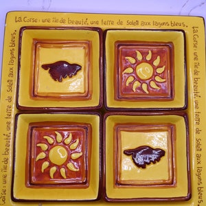 Pode incluir: Um azulejo de cerâmica com quatro quadrados, cada um com um design diferente. O quadrado superior esquerdo tem uma silhueta marrom de um pássaro, o quadrado superior direito tem um sol amarelo com raios vermelhos, o quadrado inferior esquerdo tem um sol amarelo com raios vermelhos, e o quadrado inferior direito tem uma silhueta marrom de um pássaro. O azulejo é decorado com um esquema de cores amarelo, laranja e marrom. O azulejo tem o texto "La Corse: une île de beauté, une terre de Soleil aux lagons bleus." escrito nele.