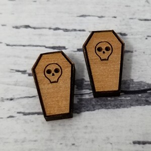 May include: Two wooden coffin-shaped earrings with a skull design. The earrings are a light brown color with a dark brown outline.