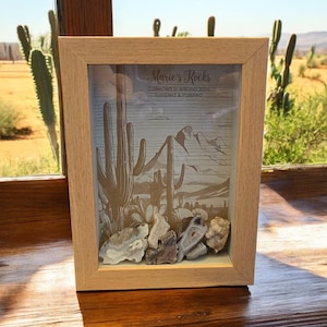 Peut inclure: Un cadre en bois avec une face en verre présente une collection de roches. Les roches sont disposées sur un fond imprimé représentant un paysage désertique avec des cactus et des montagnes. Le texte "Marie's Rocks Collected in Arizona 2014 Tumbled & Polished" est imprimé sur le fond.