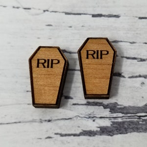 May include: Two wooden coffin-shaped ornaments. Each ornament is light brown with a dark brown border and the letters "RIP" engraved on the front. The ornaments are likely for Halloween or a similar themed event.