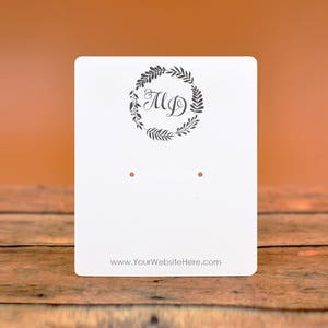 May include: White card stock earring display card with two holes for earrings. The card has a wreath with the initials "M.D." and the text "www.YourWebsiteHere.com".