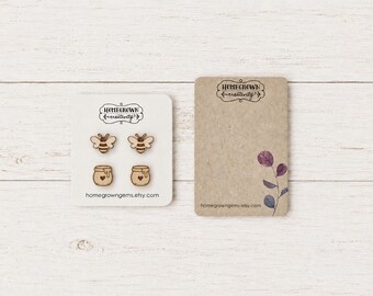 May include: Two pairs of light-coloured wooden stud earrings featuring laser-engraved bee and honey pot designs. The earrings are displayed on a white card labelled Homegrown Creativity with the URL homegrowngems.etsy.com printed at the bottom.