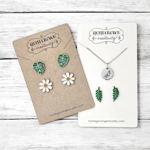 May include: A jewelry set featuring two display cards labeled Homegrown Creativity. One card holds two green monstera leaf stud earrings and two white daisy stud earrings. The other card holds a silver necklace with a leaf charm and two fern earrings.