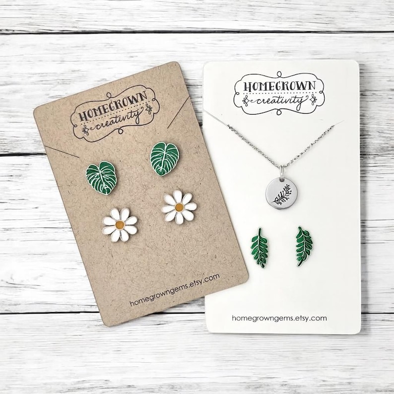 May include: A jewelry set featuring two display cards labeled Homegrown Creativity. One card holds two green monstera leaf stud earrings and two white daisy stud earrings. The other card holds a silver necklace with a leaf charm and two fern earrings.