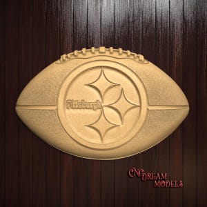 Puede incluir: Un balón de fútbol de madera con el logotipo de los Pittsburgh Steelers y la palabra "Pittsburgh" tallada en el lateral. El balón está sobre un fondo de madera oscura.