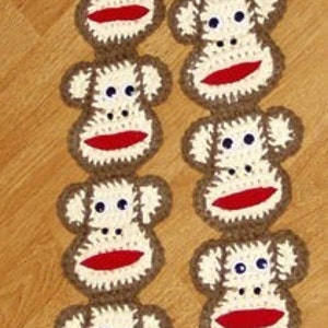 May include: A long garland of crocheted monkey faces in brown and white with red lips. The monkeys are arranged in two rows, with each row alternating between facing left and right.