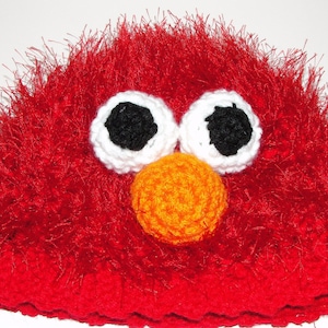 May include: Red fuzzy crocheted hat with white and black eyes and an orange nose. The hat is designed to look like Elmo from Sesame Street.