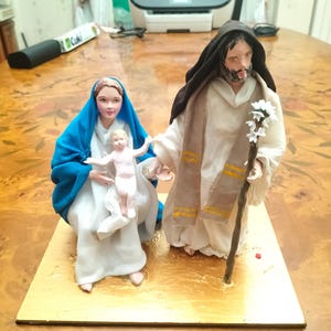 Op de afbeelding: Een handgemaakte kerststalfiguurtjesset met Maria, Jozef en baby Jezus. Maria draagt een blauwe mantel en een witte jurk, Jozef draagt een bruine mantel met een gouden rand en baby Jezus draagt een witte mantel. De figuurtjes staan op een houten basis.