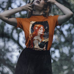 Peut inclure: Une femme portant un t-shirt orange brûlé avec un motif représentant une femme aux cheveux roux tenant un chat noir. Le motif comprend également un croissant de lune et des étoiles. La femme porte une jupe noire et a des ongles noirs.