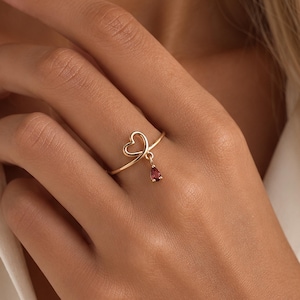 May include: A gold-toned ring featuring a delicate open heart design on the band. A small, teardrop-shaped dark red gemstone charm hangs from the bottom of the heart, creating a subtle dangling effect on this thin, polished metal finger ring.