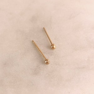 Op de afbeelding: Een paar gouden oorbelletjes met een bolletje. De oorbelletjes zijn simpel en minimalistisch, met een klein gouden bolletje aan het einde van een dunne gouden pen.