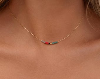 May include: A delicate gold chain necklace featuring a small, horizontal row of five tiny cylindrical beads in grey, red, black, gold, and turquoise colours centred along the thin metallic strand resting against the neck.