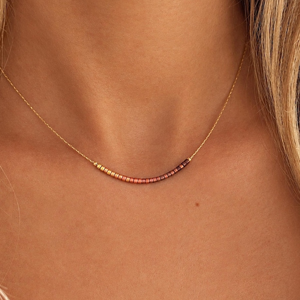 Op de afbeelding: Een delicate gouden ketting met een gebogen staafhanger bestaande uit kleine, veelkleurige kralen die in een verloop van lichtgeel naar oranje, rood en donkerpaars overgaan, rustend tegen de basis van de nek.