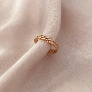 Peut inclure: Une bague dorée avec un motif tressé. La bague est présentée sur un tissu rose pâle et doux. Le motif complexe de la bague est visible, mettant en valeur son savoir-faire délicat. Cette bague est un bijou.