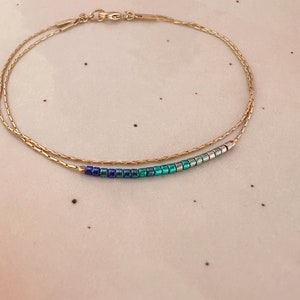 Könnte beinhalten: Ein zartes Armband aus goldener Kette mit einer Reihe von schillernden blauen und grünen Perlen.