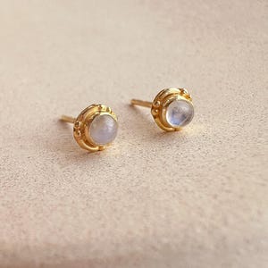 Może przedstawiać: Para złotych kolczyków sztyftów z okrągłym, jasno zabarwionym kamieniem pośrodku. Kolczyki mają dekoracyjne, teksturowane obramowanie. Sztyfty są prezentowane na neutralnej powierzchni.