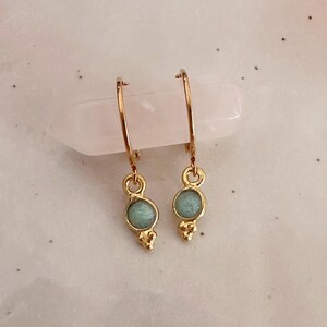Peut inclure: Une paire de boucles d'oreilles créoles en or avec un petit pendentif rond en pierre vert clair sur chaque créole.