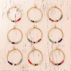 May include: Nine pairs of gold hoop earrings, each featuring a row of colourful beads. The beads include shades of red, blue, green, pink, and black, with gold beads. The earrings are displayed on a white, distressed wooden surface.