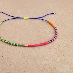Puede incluir: Un brazalete de cuentas de colores con cuentas amarillas, azules, rosas y naranjas. El brazalete está hecho con un cordón azul y tiene un nudo deslizante para ajustar el tamaño.