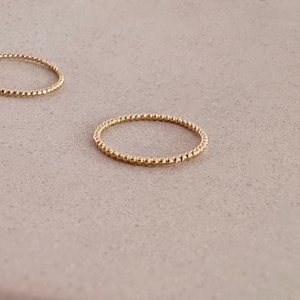 Puede incluir: Dos delicados anillos dorados con un diseño de cuentas. Los anillos son circulares y se encuentran sobre una superficie de tono neutro. Los anillos están hechos de pequeñas cuentas espaciadas uniformemente, creando una apariencia texturizada.