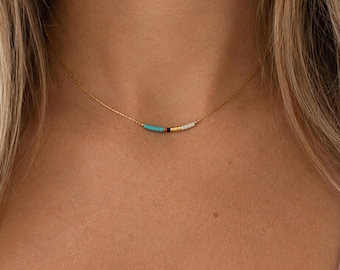 May include: A delicate gold chain necklace featuring a small horizontal bar of tiny beads in turquoise, black, gold, and white. The necklace rests against the neck, showcasing a minimalist design with a thin metallic chain and a central bead accent.