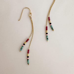 Op de afbeelding: Een paar goudkleurige oorbellen met dunne kettinkjes die aan gebogen oorhaakjes hangen. Elke oorbel heeft twee bungelende strengen versierd met kleine, cilindervormige kralen in afwisselende kleuren van turkoois, zwart, roze en donkerrood.