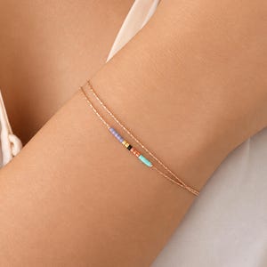 Könnte beinhalten: Ein zartes, zweireihiges Armband aus roségoldener Kette mit einem kleinen Abschnitt aus winzigen zylindrischen Perlen in Helllila, Schwarz, Orange und Türkis. Das Armband besteht aus zwei dünnen, texturierten Metallketten.