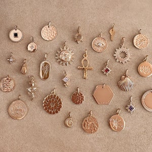 Peut inclure: Une collection de breloques dorées aux motifs variés, dont un soleil, un œil, une fleur, une étoile, une clé, une coquille et un lotus. Les breloques sont disposées sur une surface brun clair.