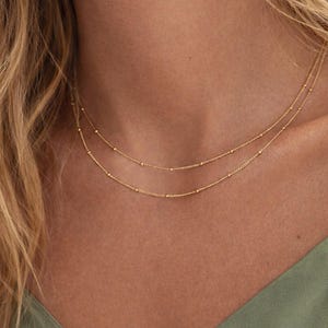 Pode incluir: Duas delicadas correntes de ouro, cada uma com pequenos detalhes de contas, são sobrepostas no pescoço de uma pessoa. As correntes contrastam com um top verde e cabelo loiro. As correntes parecem ser feitas de uma corrente fina.
