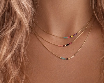 May include: A layered gold chain necklace set featuring three delicate strands. Each chain is adorned with a small, horizontal segment of tiny, multi-coloured glass beads in shades of pink, teal, black, yellow, and white.