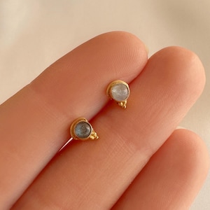 Peut inclure: Une paire de petites boucles d'oreilles à clous dorées. Chaque boucle d'oreille présente une pierre ronde de couleur claire sertie dans une lunette dorée. Les boucles d'oreilles ont un design simple et élégant, adapté au quotidien. Les pierres mesurent environ 0,6 cm de diamètre.