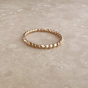 May include: A thin, circular ring crafted from polished rose gold metal. The band features a continuous, textured design composed of small, rounded, pebble-like beads linked together to form a delicate, uniform, and reflective metallic circle.